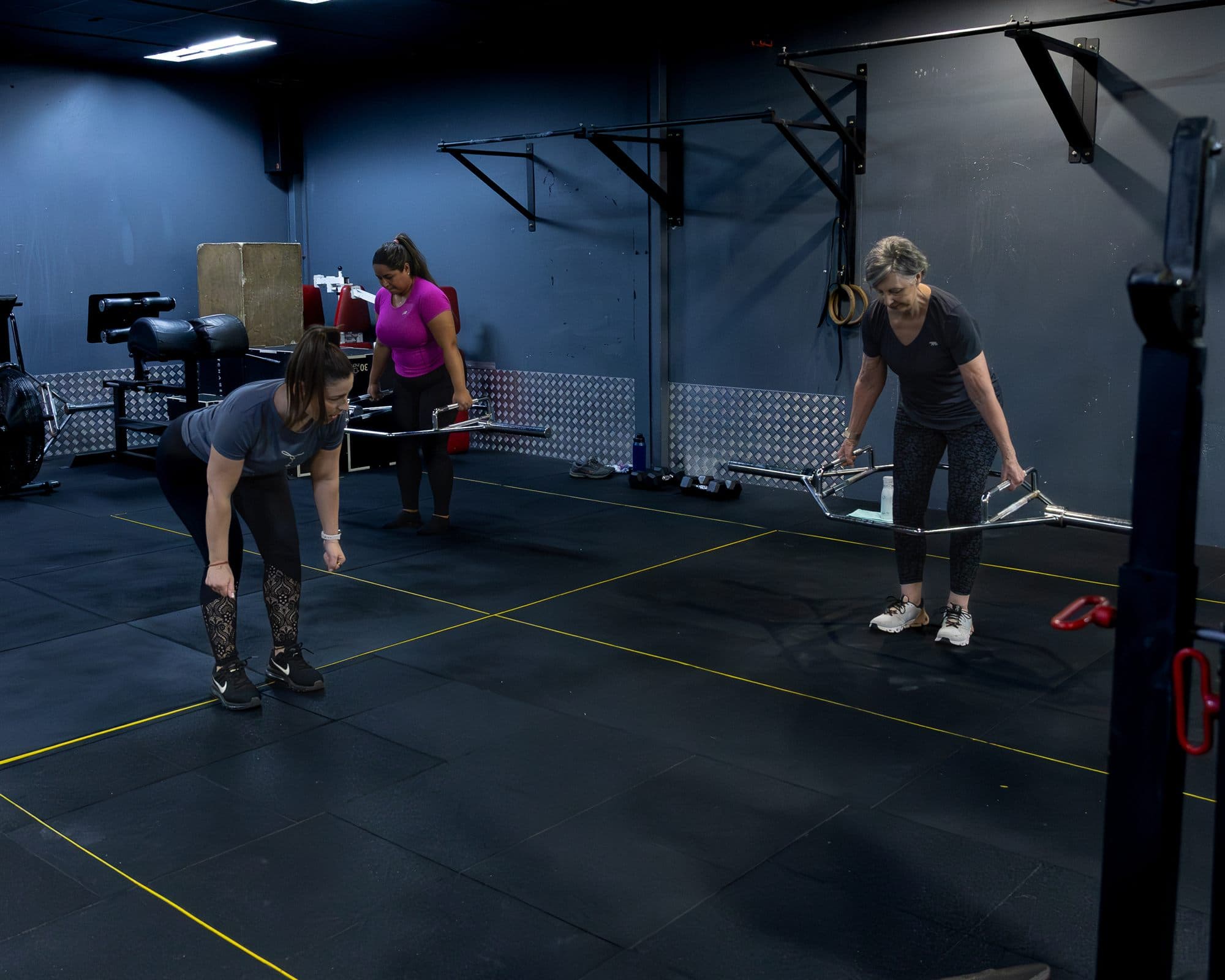 Two clients hinging at the hips over barbells while Alex watches their form from behind