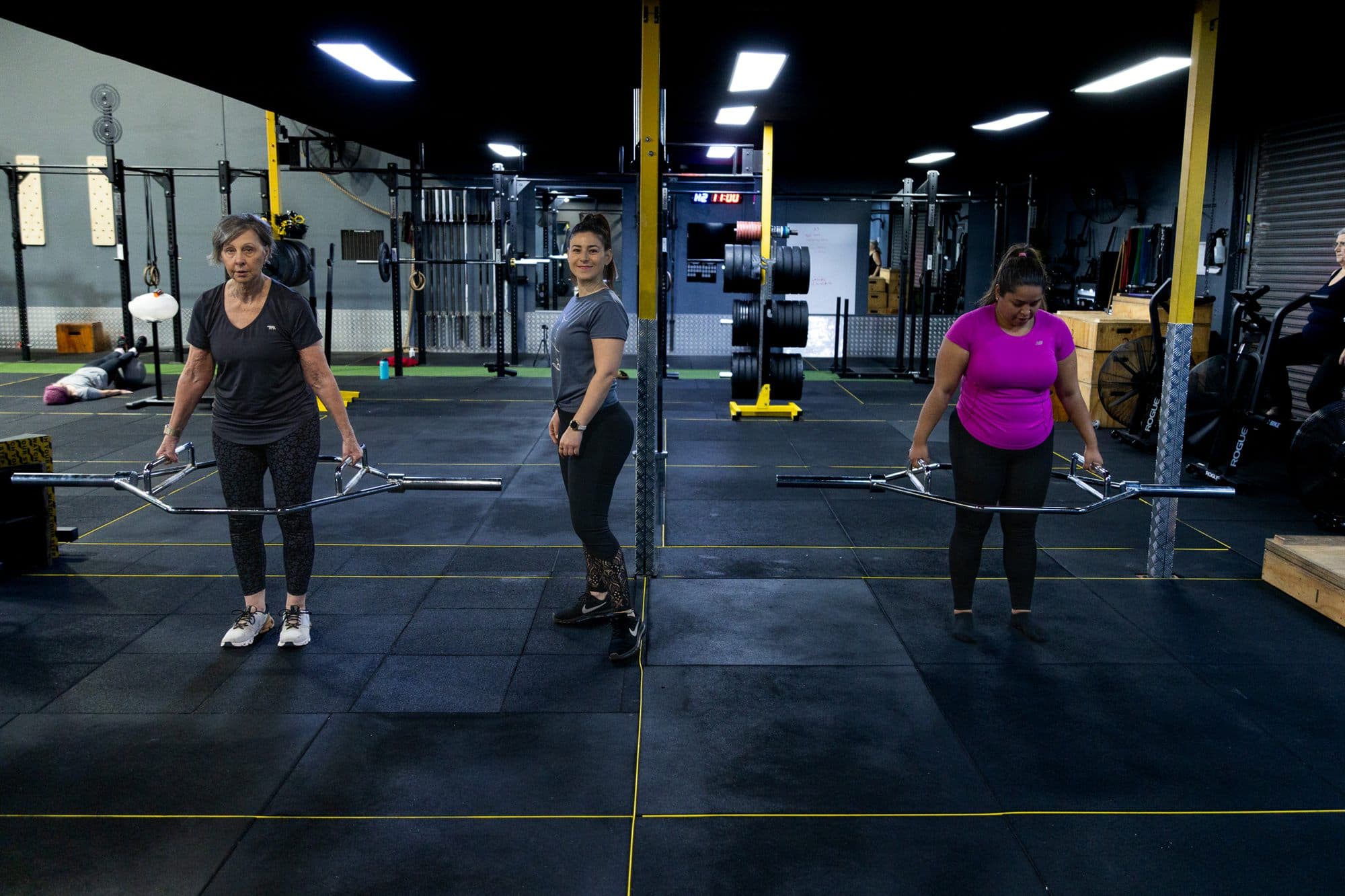 Three women spaced across the gym floor, each standing over a loaded barbell
