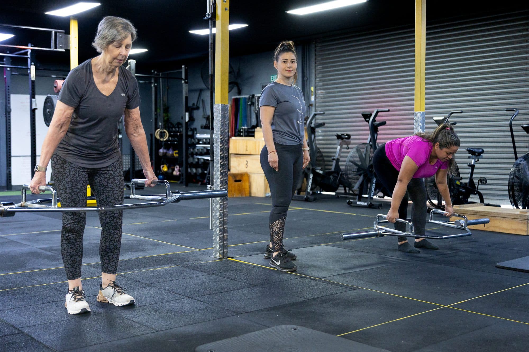 A client in her sixties holding a barbell and smiling, others training behind her